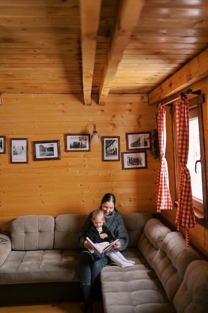 Mamãe lê um livro para uma garotinha sentada no colo no sofá de uma casa de madeira