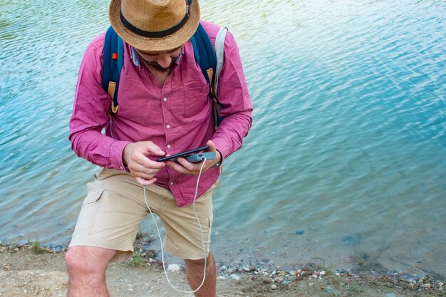 Man lädt ein Smartphone mit einem tragbaren Ladegerät auf. Ein Mann mit einer Powerbank in der Hand auf dem Hintergrund des Meeres an einem sonnigen Tag mit Wolken.