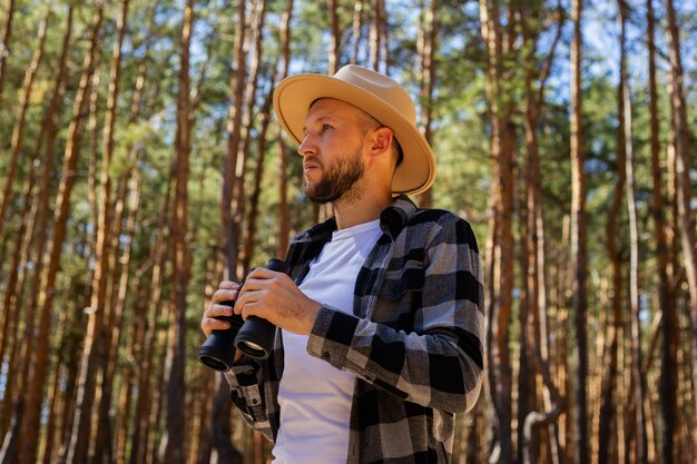 Man Tourist in einem Hut und einem karierten Hemd schaut durch Fernglas im Wald.