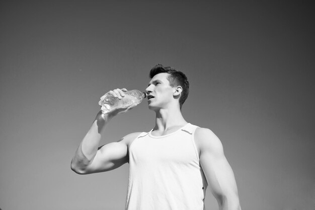 Man trinkt Wasser aus der Flasche an einem sonnigen Tag Sportler im grünen T-Shirt am blauen Himmel Bodybuilder mit muskulösen Händen Muskeln Bizeps Trizeps Sport- und Fitnesskonzept Gesunder Lebensstil und Gesundheit