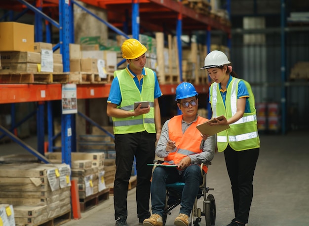 Manager, der im Rollstuhl behindert ist, Mitarbeiter, der Tablet-Check-Lagerarbeiten im Lager verwendet Arbeiter, die gut sichtbare Kleidung und Schutzhelmhelm tragen und die Anzahl der Warenkisten für die Lieferung überprüfen