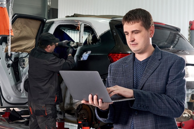 Manager, der Laptop hält, während in der Nähe von Auto in Autowerkstatt mit Mechaniker, der Auto auf Hintergrund repariert