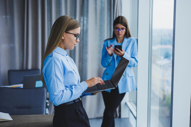 Manager-Frau in klassischer Brille, die während der Arbeitszeit im Büro lächelt positive europäische Frau im blauen Hemd Schreibtisch mit Laptop-Fernarbeit Ein Kollege ist im Hintergrund selektiver Fokus