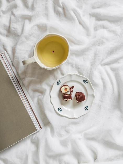 Una mañana acogedora en la cama una taza de té verde un caramelo de chocolate una revista en sábanas blancas vista de arriba