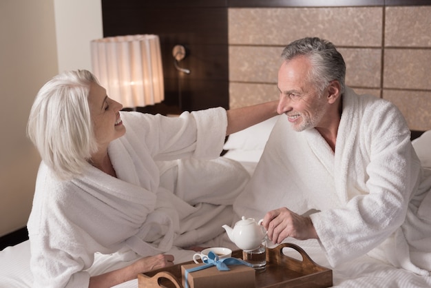 Mañana. Alegre feliz pareja de ancianos acostada en la cama y desayunando mientras expresa alegría