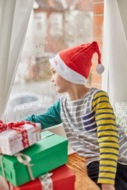 Mañana de Navidad en una casa familiar Un niño con un gorro de Papá Noel mirando por la ventana de un dormitorio