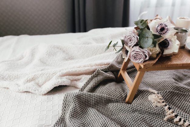 Mañana romántica. Mesa de centro de madera con flores en la cama con cuadros, taza de café, flores y velas. Rosas lilas con eucaliptos y anémonas. Tonos grises interiores.