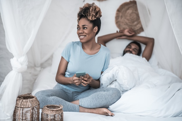 Mañana tranquila. Linda joven afroamericana en ropa de casa sentada con smartphone en la cama y esposo acostado debajo de la manta