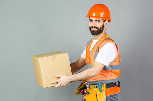 Un manbuilder en un casco naranja con una caja de cartón en sus manos Sobre un fondo gris