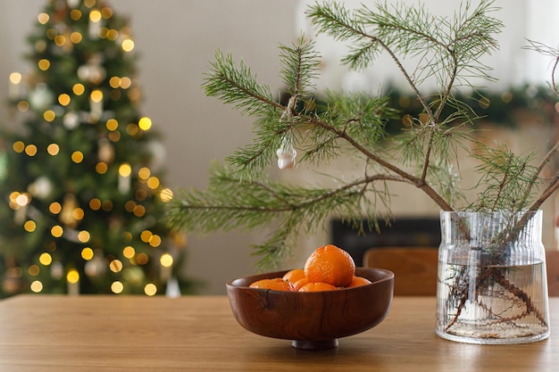 Mandarinas frescas en un tazón de madera y rama de abeto con adorno de estrella contra un elegante árbol de Navidad decorado con luces doradas Vacaciones de invierno atmosféricas en casa Feliz Navidad