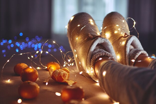 Mandarinas maduras, guirnalda de luces de Navidad blancas cálidas y piernas femeninas vistiendo cálidas y suaves zapatillas de invierno en casa acogedora en la víspera de Navidad