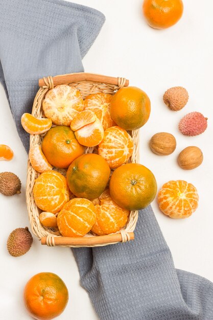 Mandarinen im Weidenkorb auf grauer Serviette. Litschi-Beeren und Walnüsse auf dem Tisch. Weißer Hintergrund. Flach liegen