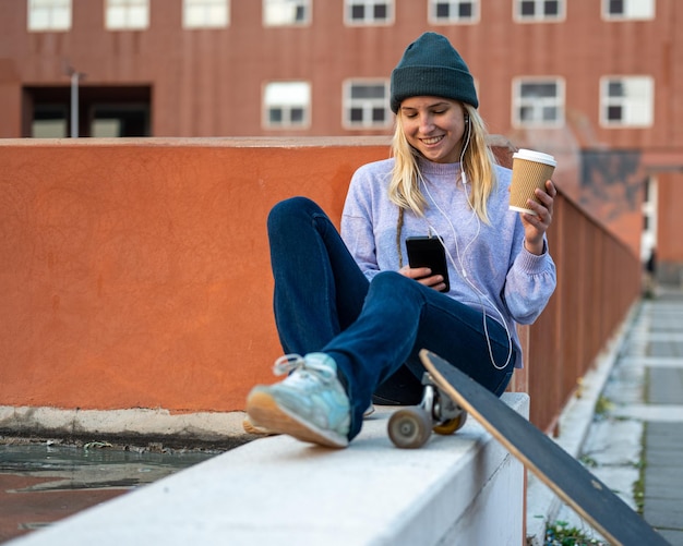 Maneira sustentável de viver jovem bebendo de uma xícara de café reciclável patinadora ouvindo música com transporte ecológico de longboard para smartphone