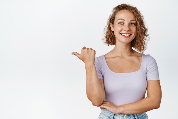 De esta manera Atractiva chica rubia señalando con el dedo a la izquierda y sonriendo mostrando un anuncio anunciando algo de pie sobre fondo blanco.