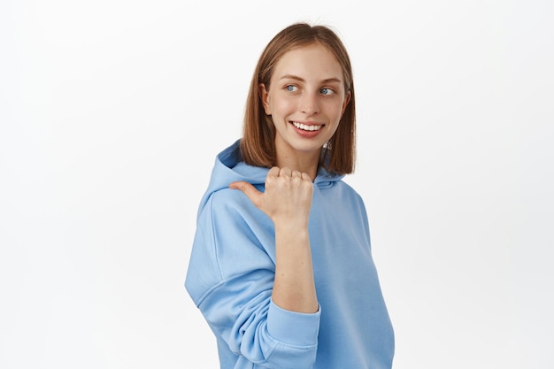 De esa manera. Chica rubia sonriente con capucha azul apuntando y mirando a la izquierda, mostrando un banner promocional de venta, texto promocional de descuento, de pie contra un fondo blanco. copia espacio