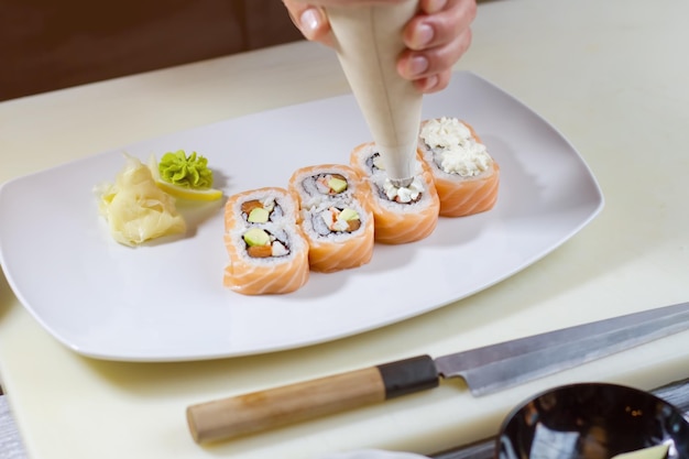 Manga pastelera sobre rollos de sushi. Herramienta pone queso crema. Plato blanco con rollos de uramaki. Últimos pasos para preparar la cena.