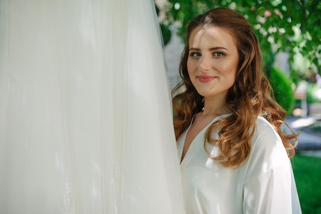 Manhã da noiva linda perto do vestido de noiva Noiva em uma túnica branca brinca com seu véu Noiva penteado noiva se veste em casa com uma túnica