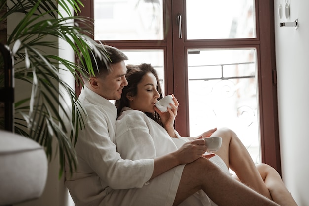 Foto manhã de jovem casal feliz em roupões de banho brancos em casa, sentado na janela, bebendo café e um abraço.