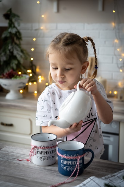 Manhã de Natal, linda menina sorridente de pijama derrama o chá do bule na xícara na mesa da cozinha.