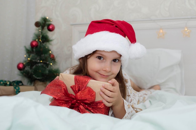 Manhã de Natal, uma menina de pijama na cama se alegra com os presentes no fundo de uma árvore de Natal. Uma criança feliz e sorridente abre um presente de Ano Novo em casa. O conceito de férias, Natal