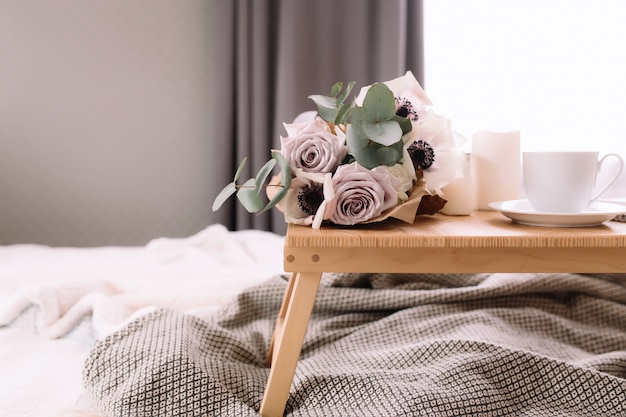 Foto manhã romântica. mesa de centro de madeira com flores na cama com manta, xícara de café, flores e velas. rosas lilases com eucalipto e anêmonas. tons de cinza interiores.