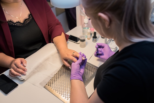 Foto manicura de hardware en un salón de belleza moderno. la cosmetóloga con una máscara facial rosa realiza el procedimiento cosmético para el cliente rubio, el lugar de trabajo profesional en segundo plano.