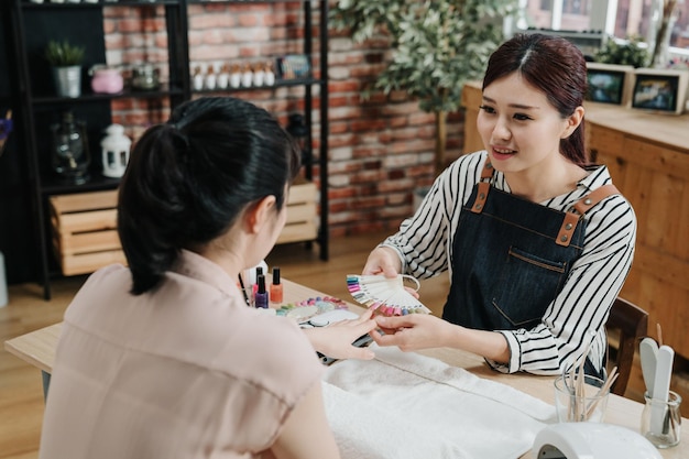 Manicura de verano y muestras de color de uñas. Maestra manicurista ayudando al cliente a elegir el color del esmalte de uñas de la paleta con el dedo de la dama en el salón de belleza de uñas. esteticista sonriente escuchar cliente hablar