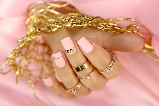 Manicure em unhas curtas com esmalte rosa branco com anéis dourados.