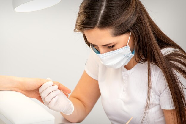 Manicurista con máscara protectora pintando uñas femeninas con esmalte de uñas en el salón de manicura.