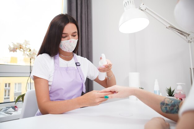 Manicurista con mascarilla, desinfectando las manos del cliente, trabajando durante la pandemia