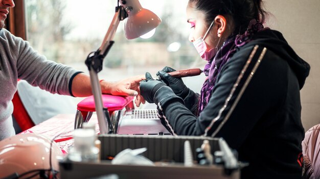 Manicurista profesional femenina haciendo manicura a un hombre latino con taladro de uñas