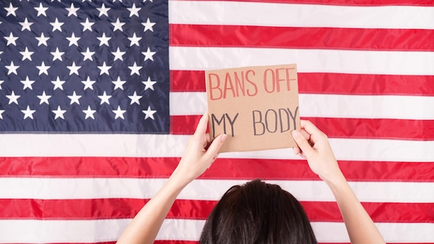 Manifestante jovem segura papelão com sinais de Bans Of My Body contra a bandeira dos EUA