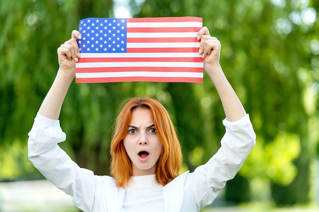 Manifestante de mujer pelirroja joven enojada posando con la bandera nacional de Estados Unidos sobre su cabeza de pie al aire libre en el parque de verano. Chica activista en manifestación con estandarte nacional.