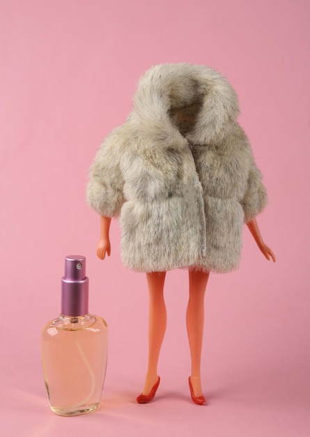 Maniquí de muñeca en abrigo de piel cálido con botella de perfume sobre fondo rosa brillante Disparo de moda minimalista Arte conceptual