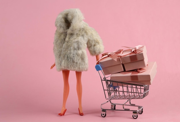 Maniquí de muñeca en abrigo de piel cálido y carrito de compras con cajas de regalo sobre fondo rosa Concepto de vacaciones minimalista Arte conceptual