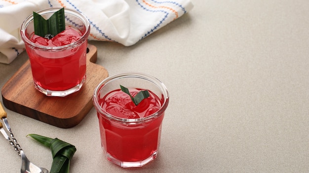 Manisan Kolang-Kaling (konservierte Zuckerpalmenfrucht) mit roter Farbe. Serviert in einem Glas mit Holzuntersetzer und Pandanblättern als Garnitur.