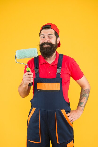 Manitas con herramienta. asistente de erector sonriente en uniforme de trabajo. ingeniero diseñador listo para pintar la pared. concepto de reparación. Trabajador de hombre barbudo con herramienta de rodillo. constructor inconformista en el fondo de la tapa amarilla.