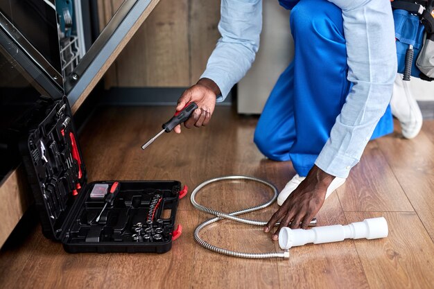 Manitas profesional irreconocible de apariencia afroamericana con un mono azul que repara el lavavajillas, usa instrumentos para abrir la caja de herramientas, en el piso de la cocina en casa. Manos de primer plano