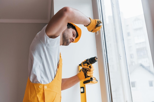 Manitas en uniforme amarillo instala una nueva ventana usando un destornillador automático Concepción de renovación de la casa