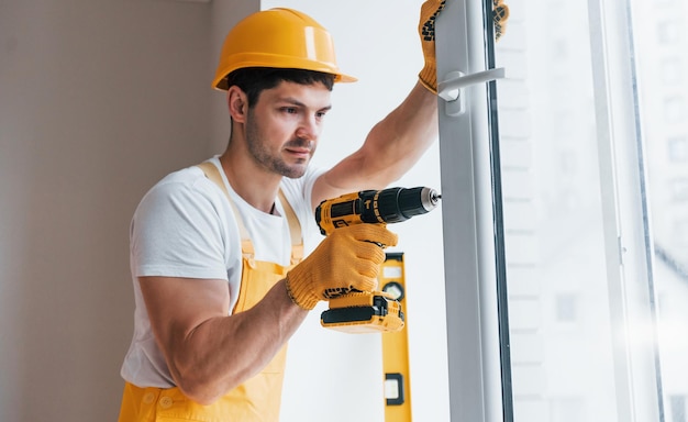 Manitas en uniforme amarillo instala una nueva ventana usando un destornillador automático Concepción de renovación de la casa