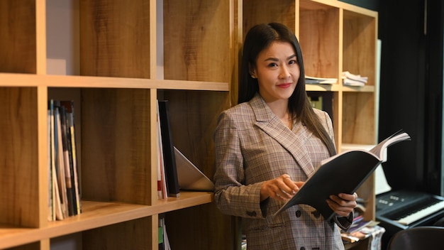 Manjedoura asiática atraente em pé perto de estantes no escritório da empresa brilhante