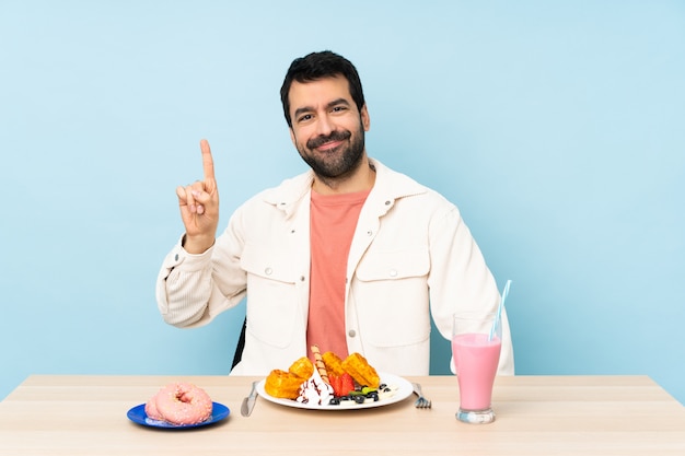 Mann an einem Tisch, der Frühstückswaffeln und einen Milchshake zeigt und einen Finger im Zeichen des Besten anhebt