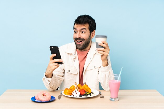 Mann an einem Tisch mit Frühstückswaffeln und einem Milchshake, der Kaffee zum Mitnehmen und ein Handy hält