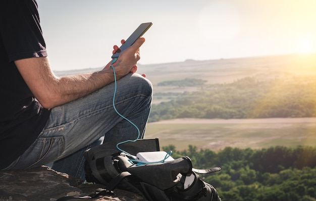 Mann auf einer Wanderung benutzt Smartphone, während er im Morgengrauen von der Powerbank auf dem Felsen auflädt