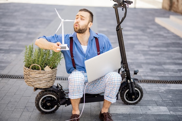 Mann bläst auf Spielzeugwindgenerator, während er mit Laptop und grüner Pflanze auf Elektroroller im Freien sitzt