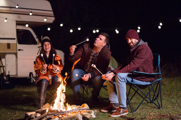 Mann bläst einen heißen Marshmallow, während Freunde in den Bergen am Lagerfeuer lachen. Retro-Wohnmobil.