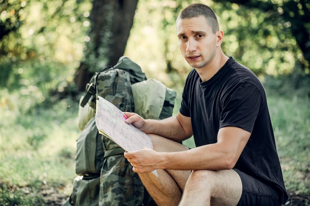 Mann, brünetter Tourist, verloren im Wald mit einer Karte, die in die Ferne schaut und versucht, den Weg zu finden. Er hat einen Rucksack, alles, was Sie für die Nacht brauchen. Suchpfade