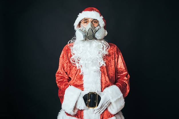 Mann, der als Weihnachtsmann mit einer Kohlefiltermaske verkleidet ist, um eine Ansteckung mit einem Virus zu vermeiden, auf schwarzem Hintergrund. Konzept von Weihnachten, Weihnachtsmann, Geschenken, Feiern.