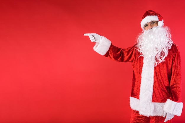 Mann, der als Weihnachtsmann verkleidet ist und mit dem Finger auf rotem Hintergrund zur Seite zeigt Weihnachtsfeiergeschenke Konsum- und Glückskonzept
