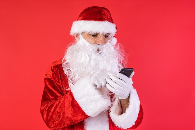 Mann, der als Weihnachtsmann verkleidet ist und sein Handy auf rotem Hintergrund konsultiert Weihnachtsfeiergeschenke Konsum- und Glückskonzept